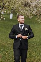 el novio vestido en un negro traje camina adelante en verde césped en naturaleza. un elegante hombre en un clásico negro traje foto