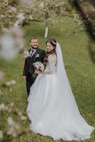 the groom and his bride are walking on the green grass in the spring garden. The bride is in a chic white dress, the groom is in a black suit photo