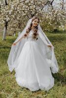 portrait of a very beautiful bride with long curly hair and wearing a white dress and long veil in a blooming spring garden. Naarechena holds her veil with her hands on a green background of grass photo