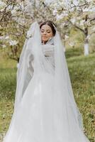 retrato de un muy hermosa novia con largo Rizado pelo y vistiendo un blanco vestir y largo velo en un floreciente primavera jardín. naarechena sostiene su velo con su manos en un verde antecedentes de césped foto