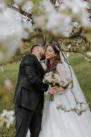 el novio Besos su novia en un blanco vestir en contra el antecedentes de floreciente arboles en un verde jardín en primavera foto