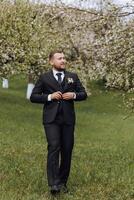 The groom dressed in a black suit walks forward on green grass in nature. An elegant man in a classic black suit photo