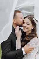 a handsome and stylish groom hugs and kisses his bride in a white dress under her veil photo