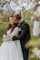 the first meeting of the bride and groom in a blooming garden. The bride in an incredible white dress with expensive hair and makeup. The groom in a black suit goes to the bride photo