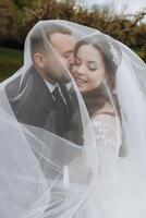 a handsome and stylish groom embraces his bride in a white dress under her veil photo