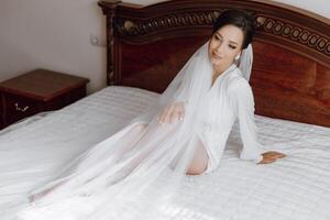 A woman in a wedding dress is sitting on a bed with a veil draped over her head photo