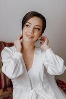 A woman in a white gown is sitting on a couch and putting on her wedding veil photo