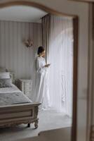 A woman in a white dress is standing in front of a bed and looking out a window photo