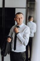 A man in a suit and tie is standing in front of a mirror, adjusting his tie photo
