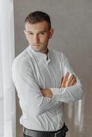 A man in a white shirt is standing in front of a window, with his arms crossed photo