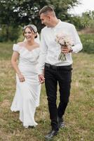 A bride and groom are walking together in a field, holding hands and smiling photo
