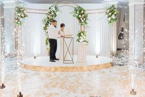 A bride and groom are cutting a cake in front of a white arch photo