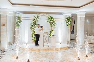 A bride and groom are cutting a cake in front of a white arch photo