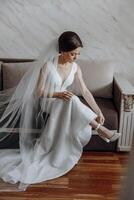 A woman in a white dress is sitting on a couch, putting on her wedding shoes photo