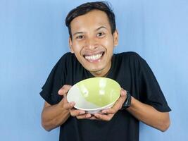 A young Asian man in a black t-shirt stands and holds an empty bowl showing the dish. photo