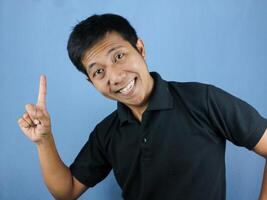 funny face expression of asian man in black shirt smiling, looking at camera while pointing up. photo
