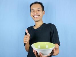 A young Asian man in a black t-shirt stands and holds an empty bowl showing the dish. photo