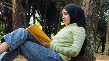 retrato de joven musulmán mujer vistiendo hijab leyendo un libro mientras propensión en contra un árbol en el parque foto