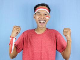 emocionado expresión joven indonesio hombre vistiendo rojo y blanco venda con apretando puño gesto. foto