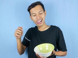 un joven asiático hombre sostiene un cuchara y vacío cuenco con el gesto de preparando a devorar el plato. foto