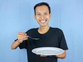 A young Asian man holds a spoon and empty plate with the gesture of preparing to devour the dish. photo