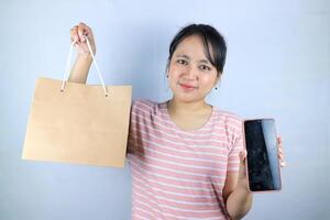 Smiling expression of Asian woman holding craft paper shopping bags and showing empty phone screen photo