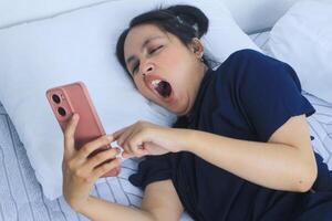 Tired beautiful Asian woman yawning in bed while holding phone. Bedtime concept photo