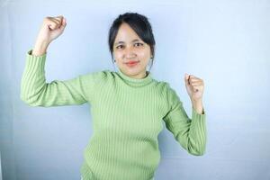hermosa asiático mujer en un verde suéter apretado puños, celebrando victoria expresando éxito foto