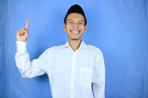 Smiling expression of muslim asian man, pointing finger an empty space. Advertising concept photo