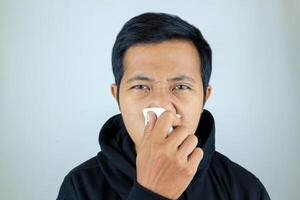 triste y infeliz asiático hombre sufrimiento desde fiebre y gripe, soplo su nariz y estornudos dentro pañuelo de papel foto