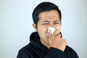 triste y infeliz asiático hombre sufrimiento desde fiebre y gripe, soplo su nariz y estornudos dentro pañuelo de papel foto