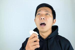 Sad and unhappy asian man suffering from fever and flu, blowing his nose and sneezing into tissue photo