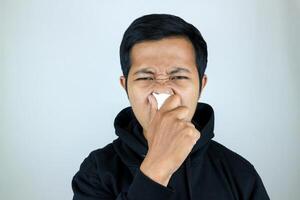 triste y infeliz asiático hombre sufrimiento desde fiebre y gripe, soplo su nariz y estornudos dentro pañuelo de papel foto