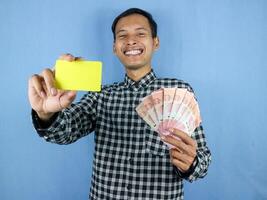 Excited expression handsome asian man hold empty card and money banknotes. Finance concept. photo