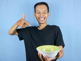 A young Asian man in a black t-shirt stands and holds an empty bowl showing the dish. photo