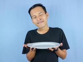 Handsome Asian man holds an empty white plate with the gesture of inhaling the dish. photo