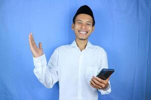 Smiling happy of muslim asian man wearing cap holding mobile phone while gesturing open hand. photo