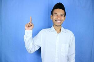 Smiling expression of muslim asian man, pointing finger an empty space. Advertising concept photo