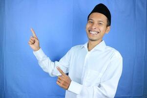 Smiling expression of muslim asian man, pointing finger an empty space. Advertising concept photo