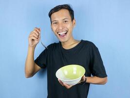 A young Asian man holds a spoon and empty bowl with the gesture of preparing to devour the dish. photo