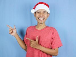 Handsome asian man in red christmas cap pointing fingers isolated over blue background. photo