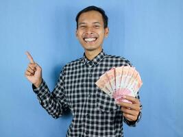 Excited expression handsome asian man hold paper money banknotes with pointing aside gesture photo