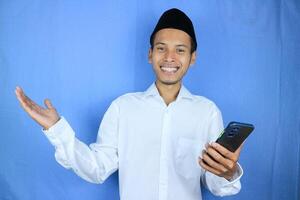 Smiling happy of muslim asian man wearing cap holding mobile phone while gesturing open hand. photo