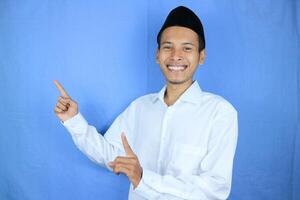 Smiling expression of muslim asian man, pointing finger an empty space. Advertising concept photo