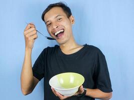 un joven asiático hombre sostiene un cuchara y vacío cuenco con el gesto de preparando a devorar el plato. foto