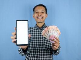 Excited expression handsome asian man hold paper money banknotes and showing blank phone screen. photo