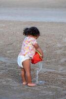 un curioso negro niño vierte arena desde un balde, absorto en jugar, un escena de inocente exploración, y niño curiosidad en temprano edad. foto