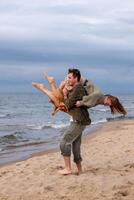 Man carrying woman piggyback on beach, both smiling, woman's legs swinging. Image captures fun, affection, ideal for themes of love or joyful moments and the happiness of weeding or engagement. photo