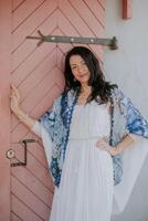 A smiling woman in a white dress and blue patterned shawl leans against a pink wooden door, exuding casual elegance and warmth. Perfect for lifestyle and fashion content. photo