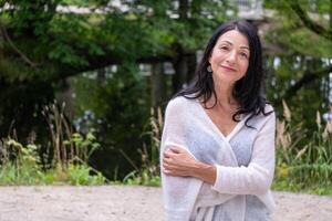 A confident mature woman in a light gray wrap smiles warmly, standing against a lush park setting, exuding self assured grace and natural elegance, as well as time in nature. photo